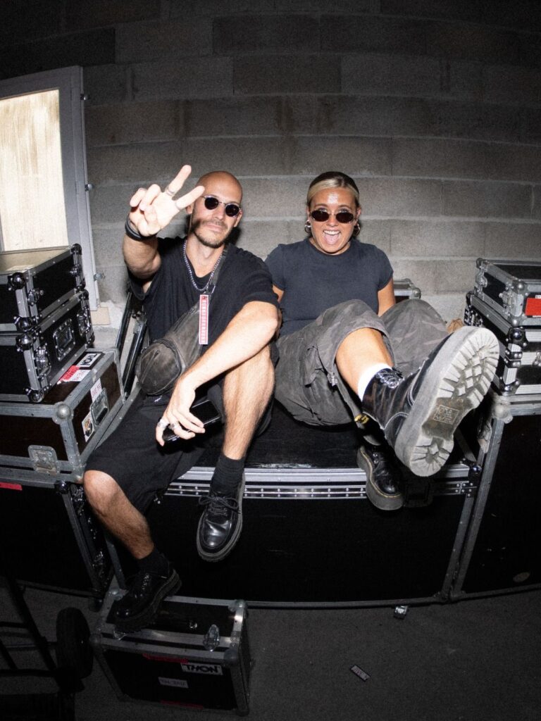 Baptiste et Tara de Jive Me en coulisses, assis sur des flight cases, Baptiste fait un signe de paix