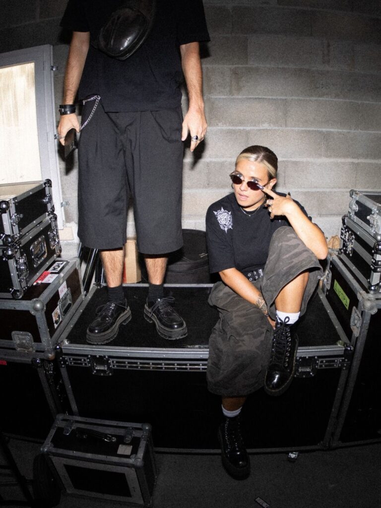 Tara de Jive Me pose en coulisses sur des flight cases, Baptiste debout derrière elle